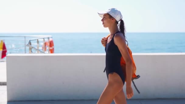 Krásná stylová sexy dáma v černém plavky, bílá čepice a oranžový batoh chůze podél zálivu u moře — Stock video