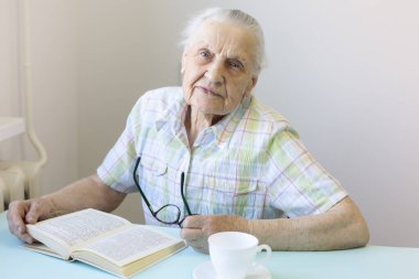 Yaşlı Kafkas Kadın Masanın yanında Oturuyor. Masa Açık Kitap, Beyaz Kupa, El Gözlüğü. Koronavirüs salgını sırasında evde kalır. Konsept - Evde Kal No COVID19..