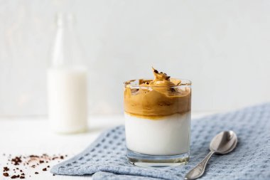 Buzlu Dalgona Kahve, modaya uygun yumuşacık kremalı kremalı kahve. Hazır kahve, şeker, sıcak su ve sütten yapılmış Kore içeceği. Gri peçeteye cam, kaşık. Beyaz arka plan. Yumuşak odaklanma. Kapat.