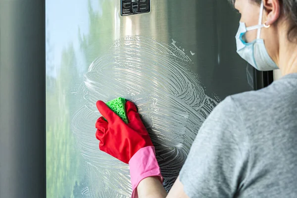 Caucasian woman in protective mask, red rubber gloves with  cleaning sponge wipes soap foam on stainless steel refrigerator door. Concept - cleaning house, cleaning service. Selective focus.