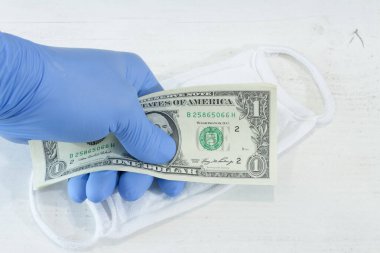 Mavi koruyucu bir eldiven içinde bir tomar banknot ve koruyucu maske tutan insan eli..