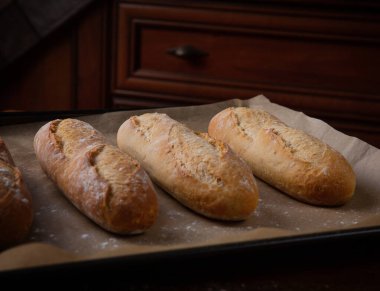 Mutfakta, kahverengi ahşap bir masanın üzerinde, fırından yeni çıkmış ekmekler fırın çarşafının üzerinde uzanıyor..