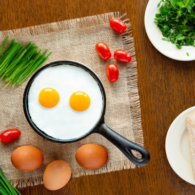 Kahverengi ahşap mutfak masasındaki çuval bezi üzerinde yumurtalı porsiyonlu bir tava var. Yanında taze vişneli domates, yeşil soğan ve taze maydanoz var.