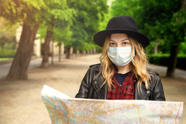 A young girl holds a city map on the street on a trip.Looks at the camera.woman with blond hair, in a leather jacket, in a hat. Protective mask on her face. A new travel concept in the new realities