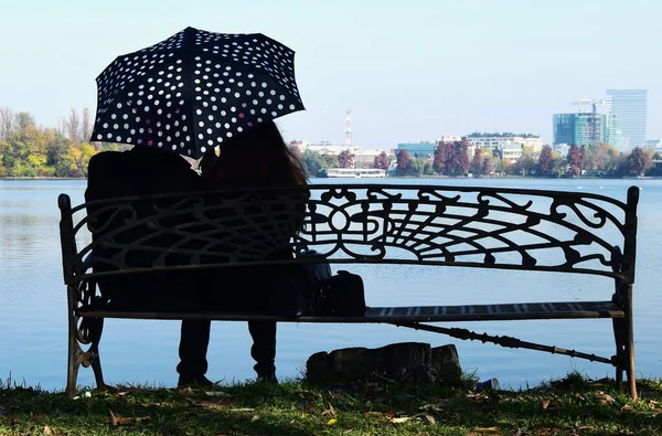 Bir şemsiyenin altında iki aşık gölün kenarındaki bankta oturuyor.
