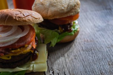 İki ev yapımı burger, ekmek, soğan, domates, et, peynir ve marul, ahşap tahtada, arkasında sos kapları var.