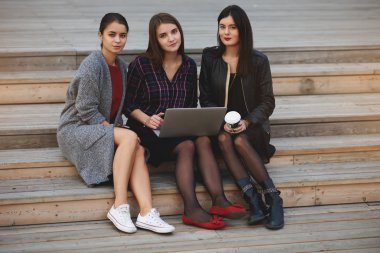 Young three hipster girls with trendy look are posing for camera after video call on net-book during their free time