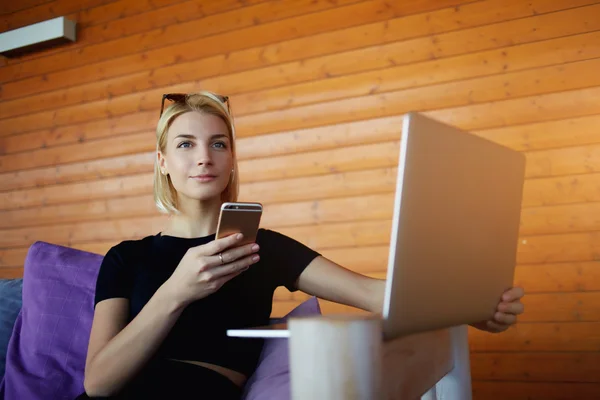 Mujer joven está pensando qué escribir es mensaje de texto en el teléfono celular, mientras que está sentado con net-book en el espacio de co-trabajo — Foto de Stock