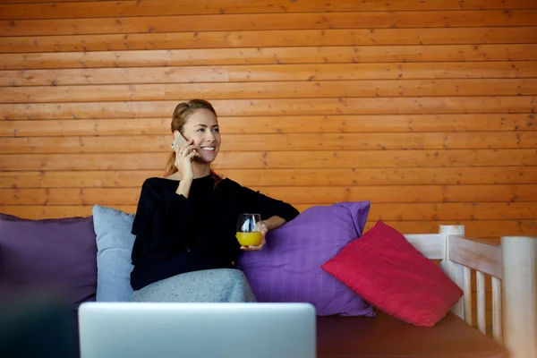 Mujer sonriente freelancer está hablando a través de un teléfono inteligente durante el descanso entre el trabajo en Internet a través de un net-book portátil . — Foto de Stock