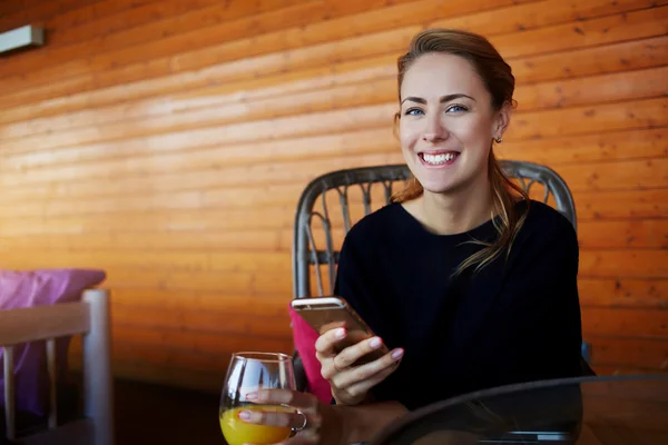 Unga glada hipster flicka med mobiltelefon i händerna tittar till kameran, tag sitter i moderna kafé interiör — Stockfoto