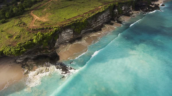 Foto aérea de drone voador de uma paisagem natural incrível com penhasco de rocha na costa arenosa. Bela água do mar com ondas para surfar na temporada de verão em Bali . — Fotografia de Stock
