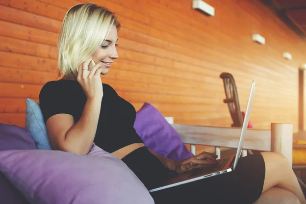 Joven hipster chica está hablando por teléfono móvil con su amigo durante el trabajo en portátil net-book, mientras está sentado en la terraza del hotel . — Foto de Stock