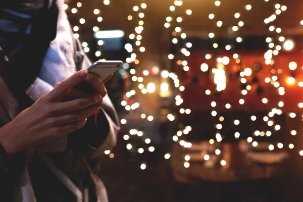 Тесно изображение молодой хипстерки девушка использует свой смартфон, в то время как стоит на улице против рождественских гирлянд огни . Стоковое Фото
