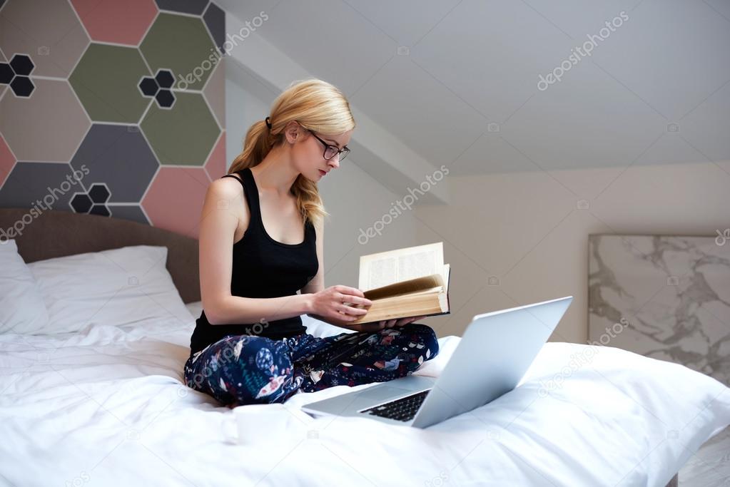 Caucasian student is using literature for learning, while is relaxing with laptop computer in her home interior