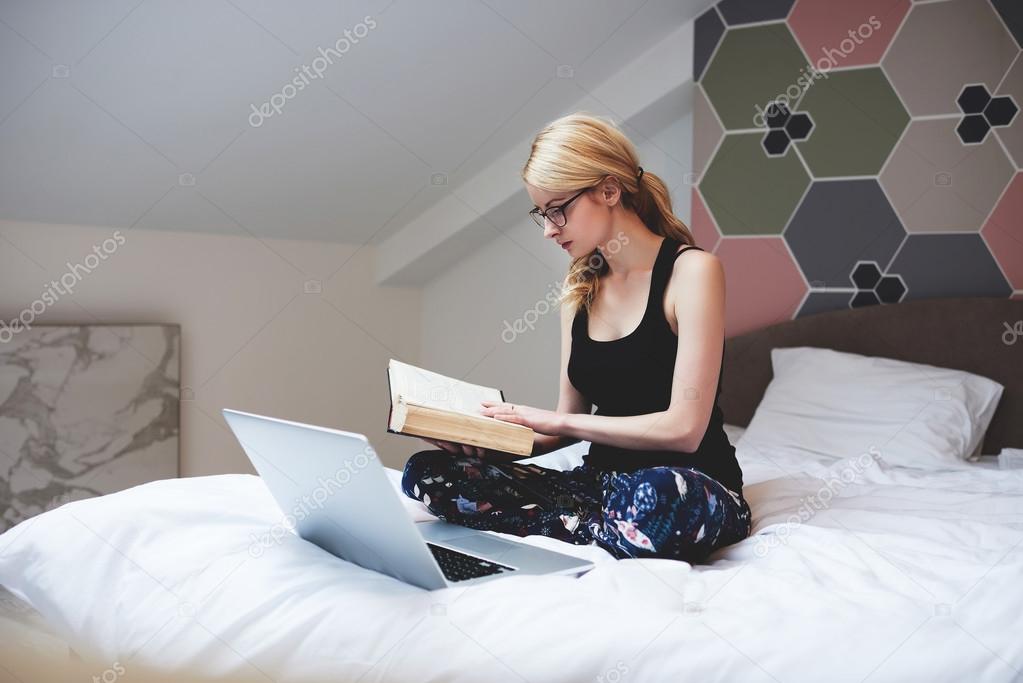 Smart student is reading book before working on coursework via laptop computer, while is sitting on comfortable bed in modern home interior.