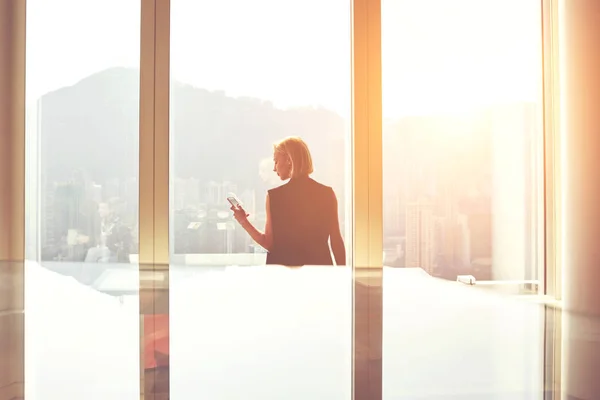 Piękna pani przy użyciu połączenia szybkie 4g i nowoczesny smartfon stoi w biurze wnętrza z widokiem na panoramę miasta — Zdjęcie stockowe