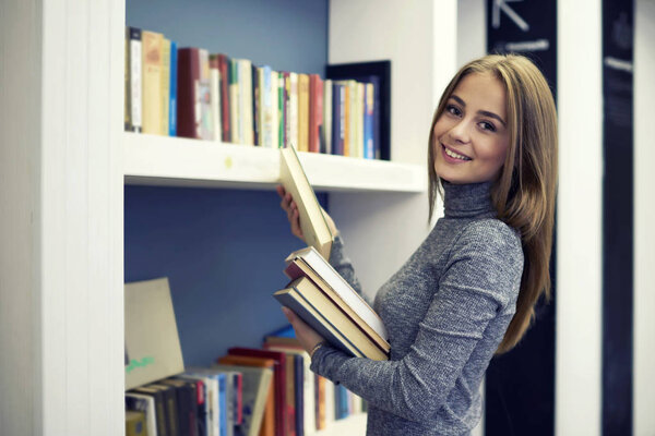 Cheerful female young professional librarian smiling while making revision of book  putting them on new bookshelves