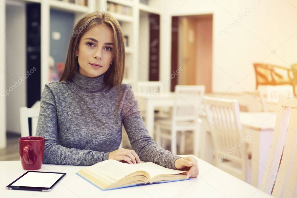 Beautiful female sitting at table in university library with free wifi zone drinking hot coffee for breakfast before starting lessons