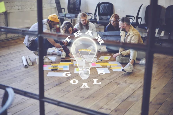 Young group of colleagues sitting on floor in modern loft interior coworking office brainstorming while analyzing information for startup project. Infographic design with conceptual teamwork goals