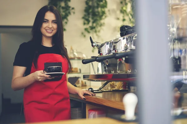 Усміхнена приваблива жіноча офіціантка насолоджується робочим процесом, що тримає чашку капучино — стокове фото