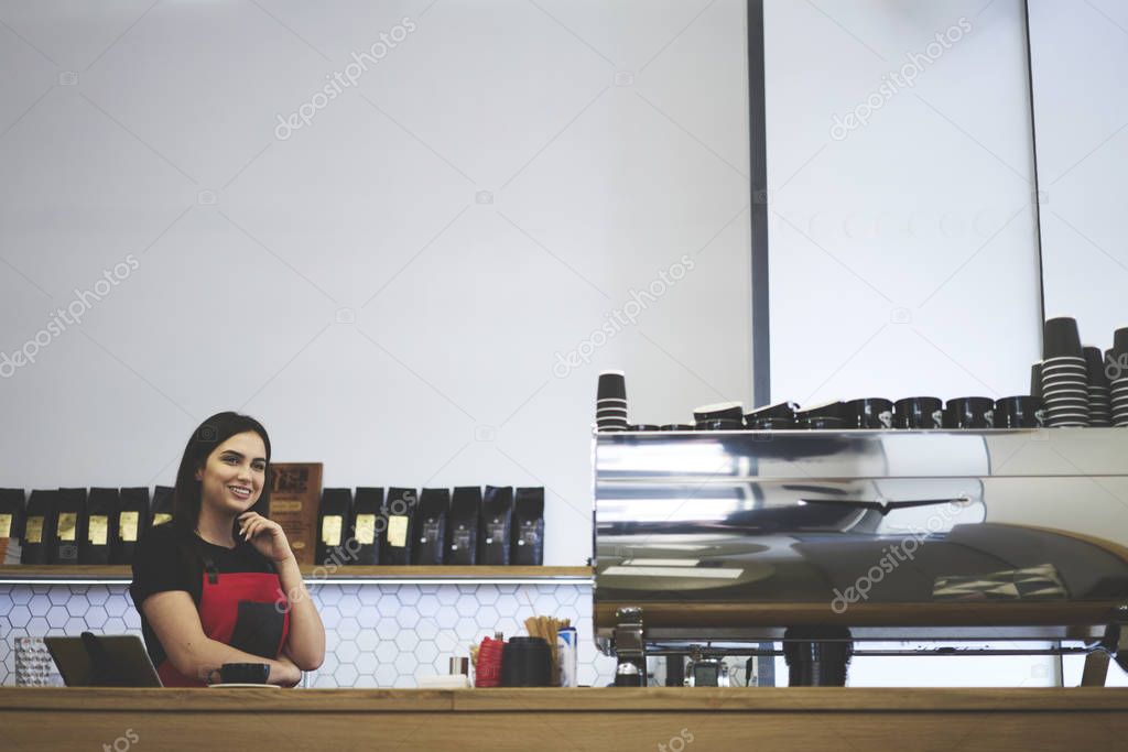 Charming female barista dreaming during working process dressed in cute uniform of coffee shops with loft interior