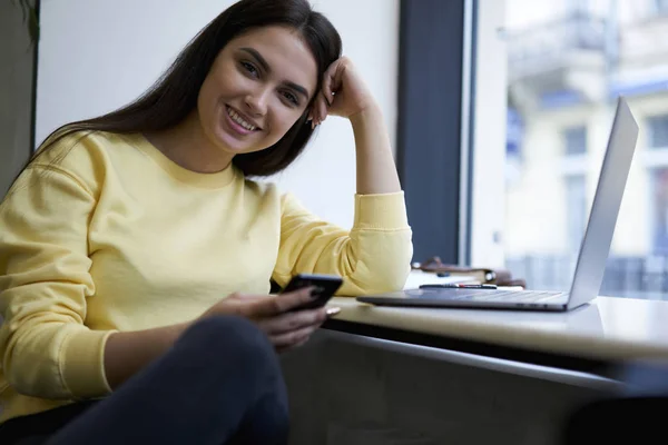 Lächelnde Cafeteria-Inhaberin, die in die Kamera schaut und das Telefon hält, um mit Kollegen zu chatten — Stockfoto