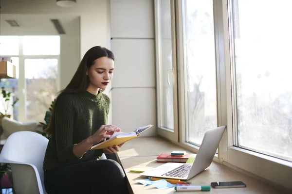 Mulher atraente inteligente lendo livro interessante e trabalhando no computador moderno no interior do espaço de co-trabalho . — Fotografia de Stock