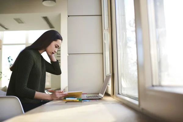 Ung kvinnelig student fokuserte på å lese bøker og søke informasjon om datamaskinen i biblioteket nær vinduet – stockfoto