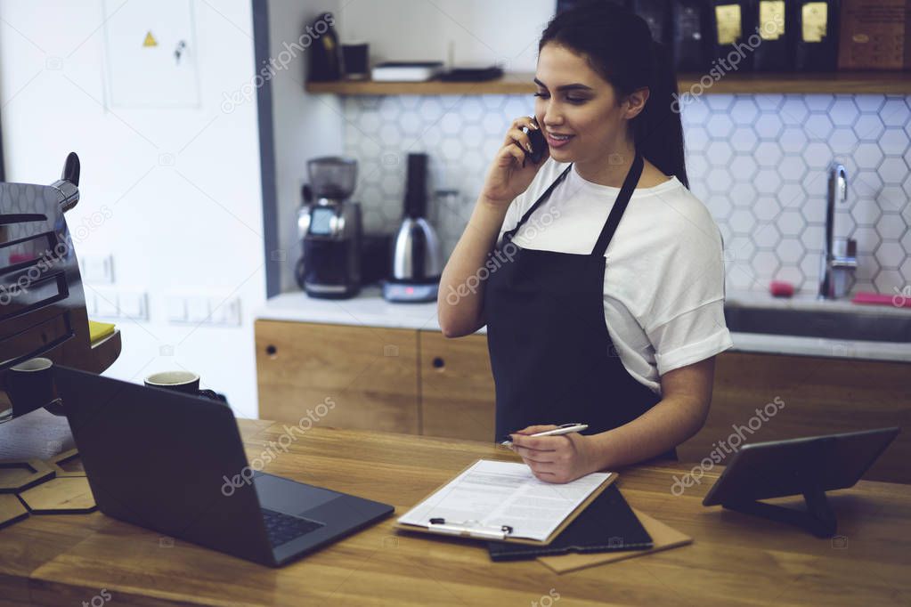 Barista taking order on cellular using paper and pen for records standing at counter with laptop