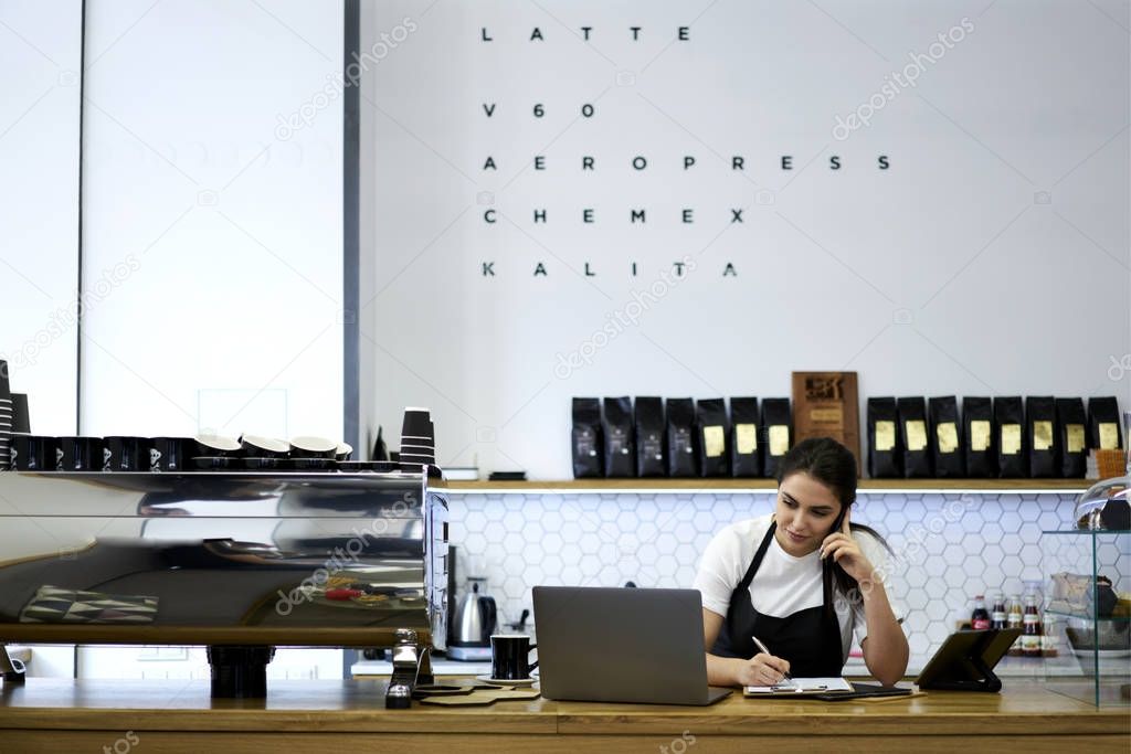 Female waitres sitting at bar with laptop while talking with administrative on smartphone