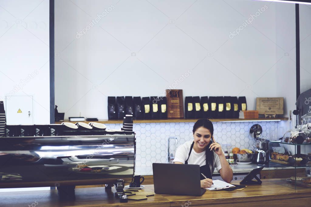 Attractive waitress talking on smartphone and making audit products at cafe indoors