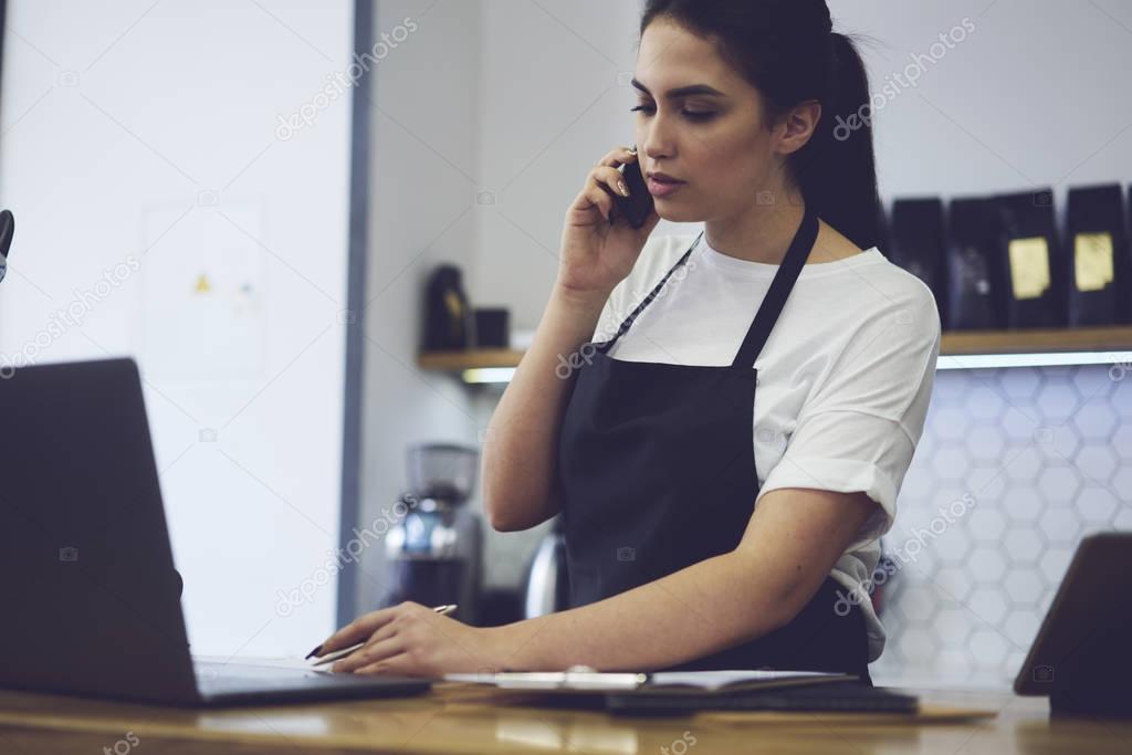 Young female owner calling to bank for checking finance documentation in cafeteria.