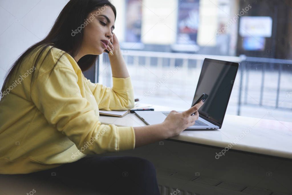 Beautiful student surfing websites on cellphone sitting in coffee shop with laptop