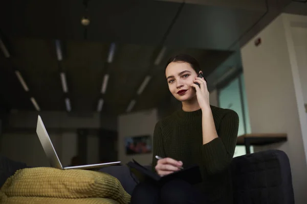 Jonge vrouw zoekt op camera tijdens het schrijven van notities in laptop zitten in de ruimte met moderne laptop coworking — Stockfoto