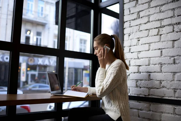 Empresaria escribiendo ideas creativas en un cuaderno mientras llama a un colega usando un teléfono inteligente moderno sentado cerca de la ventana —  Fotos de Stock