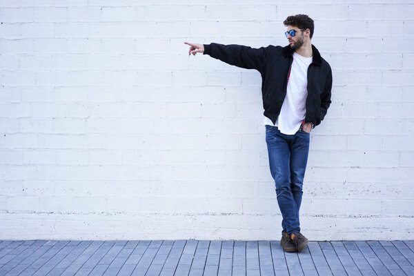 Hipster guy posing in stylish wear, pointing with finger on copy space