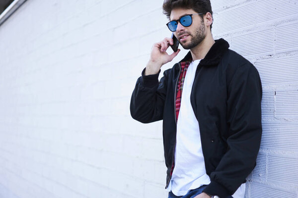 Handsome caucasian model dressed in trendy clothes and stylish sun glasses calling to best friend via cellular.