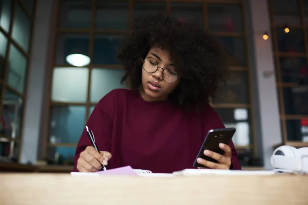 Afroamerikanische Studentin sitzt auf Uni-Campus am Tisch und blickt in Kamera mit modernem Smartphone — Stockfoto