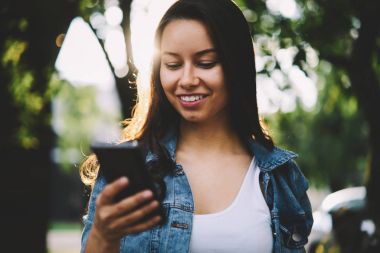 Sosyal ağlar üzerinde modern smartphone aygıt içinde dostça sohbet sırasında gülümseyen çekici güzel bayan. Kentsel ortamda neşeli genç kadın okuma sms mesaj üzerinde dijital telefon ayakta