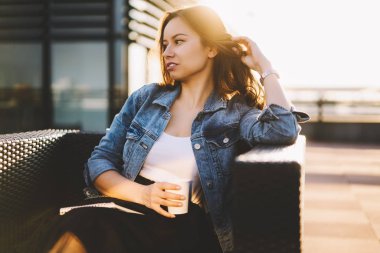 Güzel esmer işkadını uzakta açık havada eğlence vaktinizi lezzetli içecek bir bardak ile kahve dükkanında otururken arıyorum. Çekici Bayan kapuçino ile dinlenme denim ceket giymiş