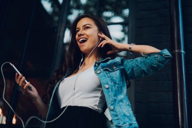 Aşağıda görünümü modern smartphone elinde tutan ve kulaklıklar radyo Web sitesinde en sevdiğiniz şarkıları dinlemek duygusal olumlu esmer kadın. Müzik keyfi denim ceket çekici genç kadın