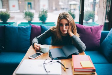 Fincan lezzetli kapuçino tutarak ve sınavları için hazırlık yaparken kameraya bakarak yorgun genç kadın portresi. Yorgun öğrenci okul sırası vasıl coworking edebiyat kitaplarında ile oturan Üstten Görünüm