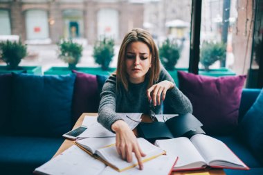 Konsantre öğrenci kahvehane iç vasıl okul sırası kağıtları ve edebiyat kitapları ile oturuyor ve eğitim projesi için yararlı bilgi arama.