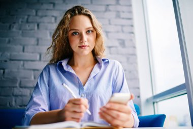 Yarım uzunluk portre fotoğraf makinesi vasıl bakarken modern cep metin bilgileri yazma kıvırcık saçlı çekici öğrenci.