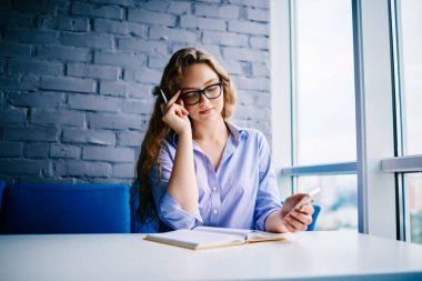 Açık kitap oturma ve uzay coworking içinde serbest zaman okuma şık gözlük akıllı öğrenci. Genç kadında akıllı telefon elinde tutan ve okuyan optik gözlük