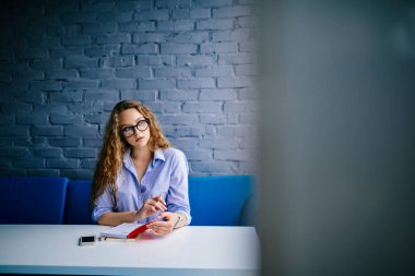 Düşünceli güzel genç kadın kıvırcık saçlı ve şık gözlük uzağa arıyorsunuz şık coworking uzayda açık kitap ile masada oturuyor. Kopyalama alanı alanı reklam metin iletinizi için