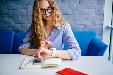 Dalgın öğrenci şık gözlük Üniversitesi iç kitaplarla masada oturan smartwatch zaman izleme.