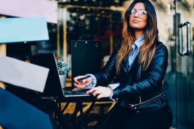 Şık genç kadın freelancer optik gözlük ve modern dizüstü bilgisayar açık havada oturma çalışma trendy deri ceket.