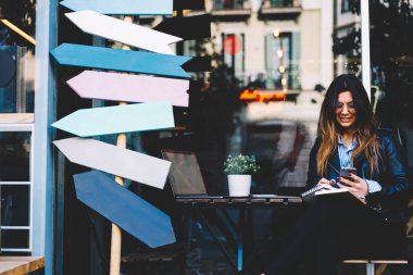 Olumlu şık hippi kız açık havada oturup kahve dükkanında netbook ile modern dijital telefon mesaj içinde not etmek aşağı yazma optik gözlük.