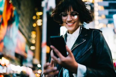 Neşeli hippi kız gece megalopolis dinleme müzik kulaklık, mutlu genç kadın illuminatio ile akşam şehirde vakit Sms okuma gezinmek için mobil uygulama kullanarak Eyewear